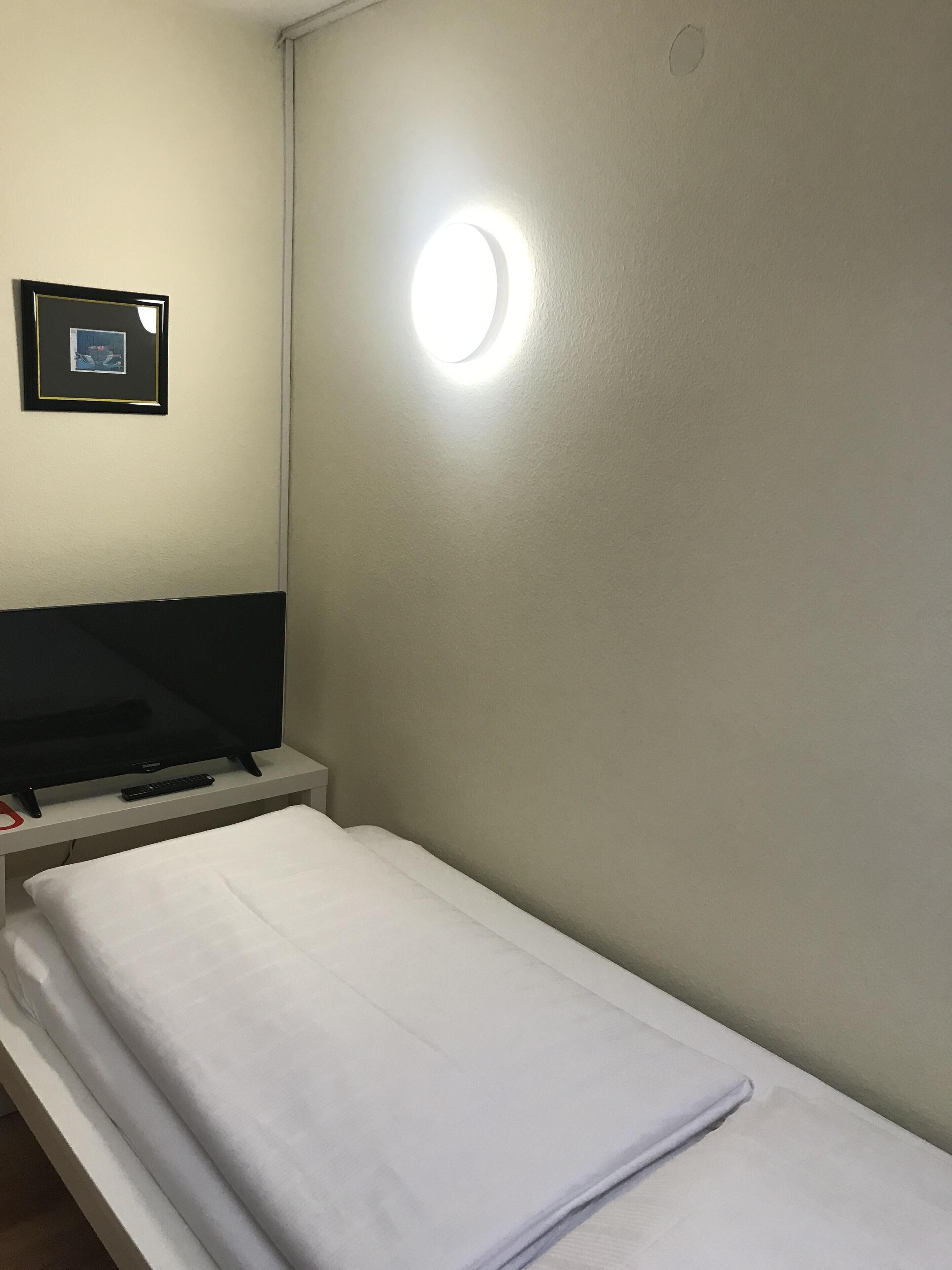 A single bed with a white mattress and a blanket, a flat-screen TV on a stand, a remote control, and a framed picture on the wall.