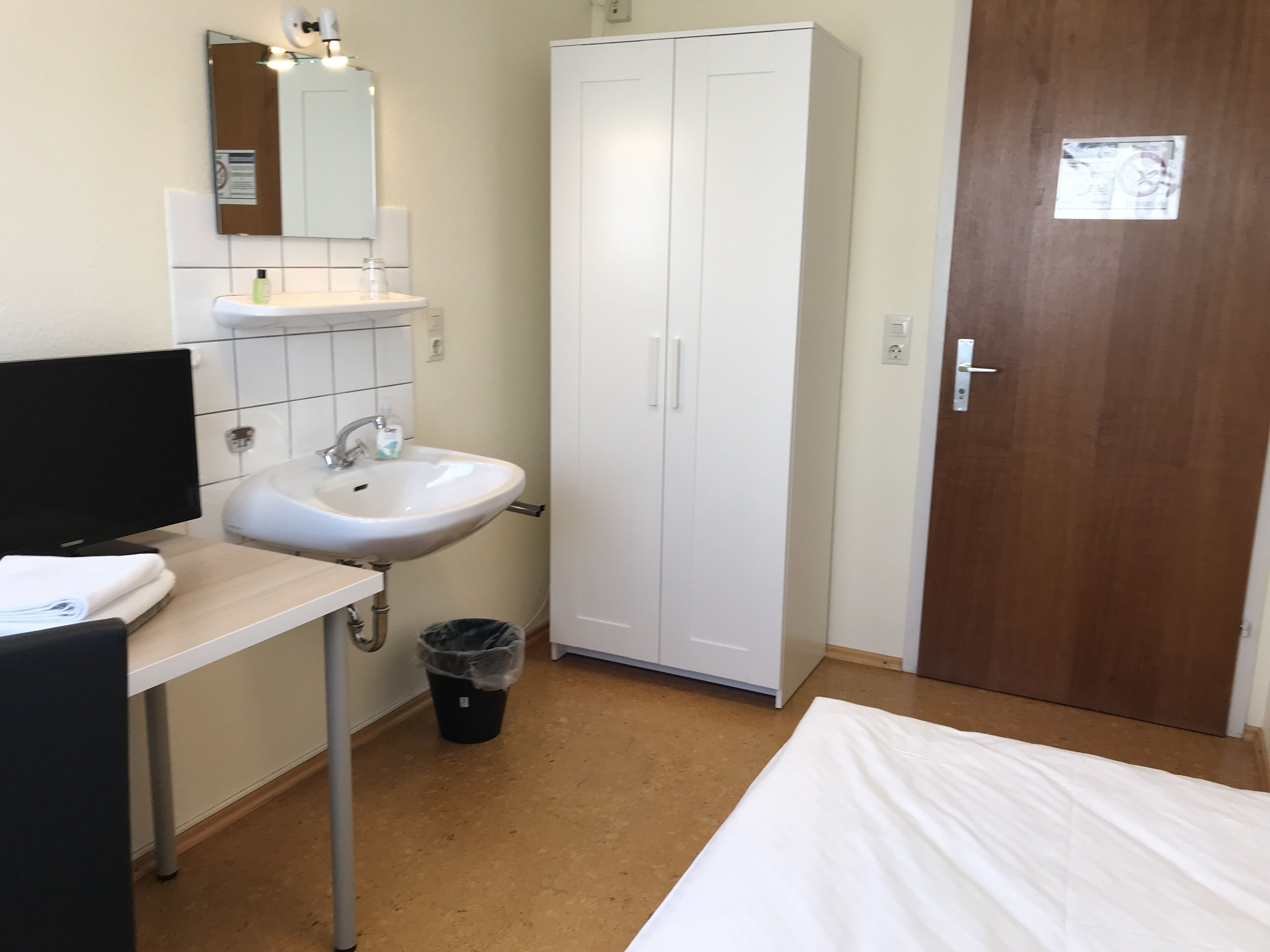 A small, clean room with a white sink, a mirror, a wardrobe, and a bed.