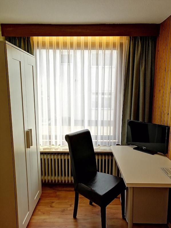 Una habitación con un escritorio blanco, una silla negra, un televisor sobre el escritorio, un radiador debajo de la ventana y un armario a la izquierda.
