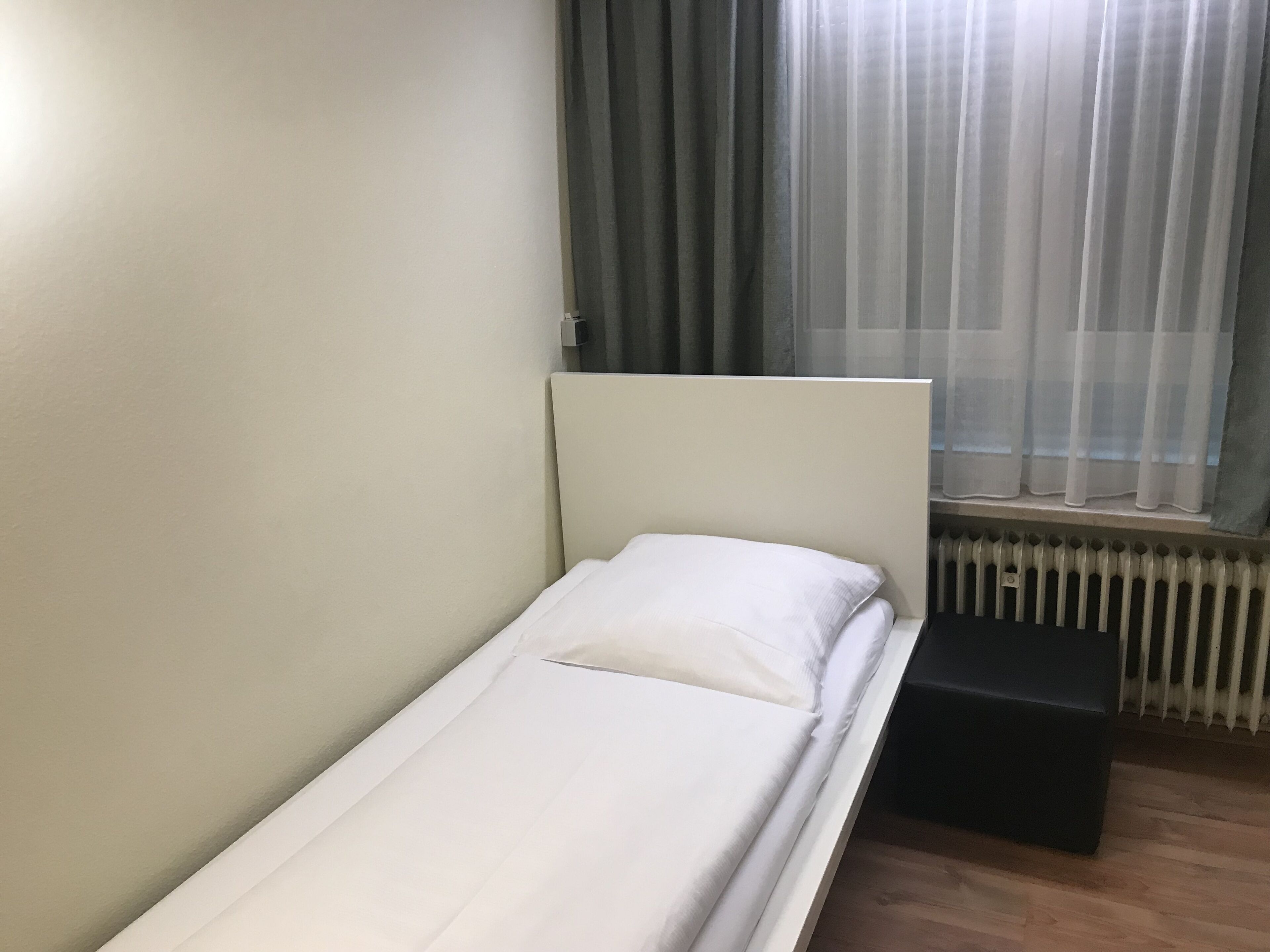 A single bed with white bedding in a room with a radiator and a window with curtains.