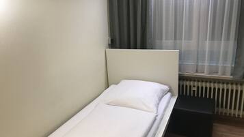 A single bed with white bedding in a room with a radiator and a window with curtains.