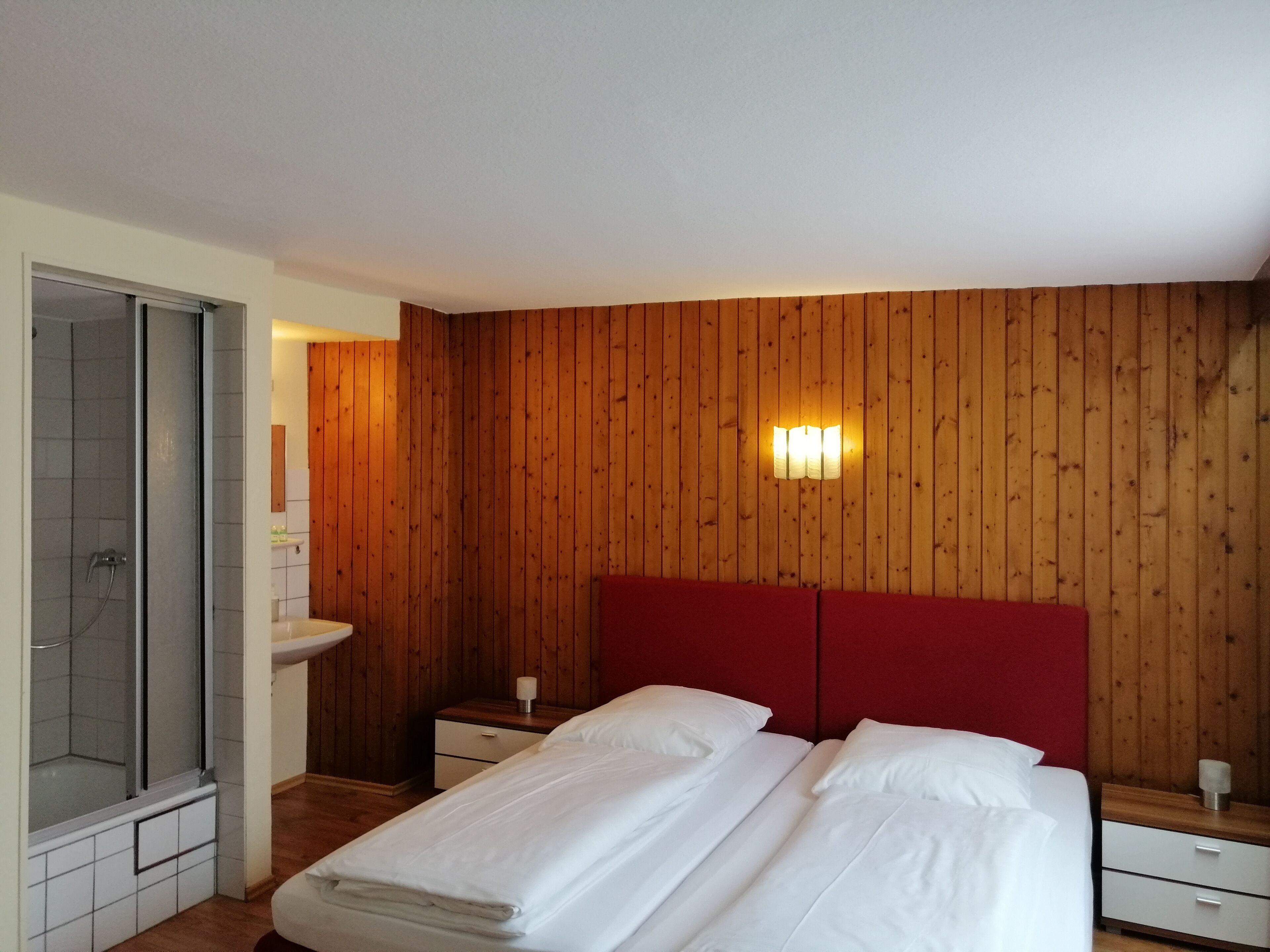 A bedroom with a wooden paneled wall, a red headboard, a double bed with white linens, a nightstand with a lamp, and a glass shower enclosure.