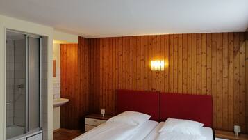 Une chambre avec un mur lambrissé en bois, une tête de lit rouge, un lit double avec du linge de lit blanc, une table de chevet avec une lampe et une cabine de douche en verre.