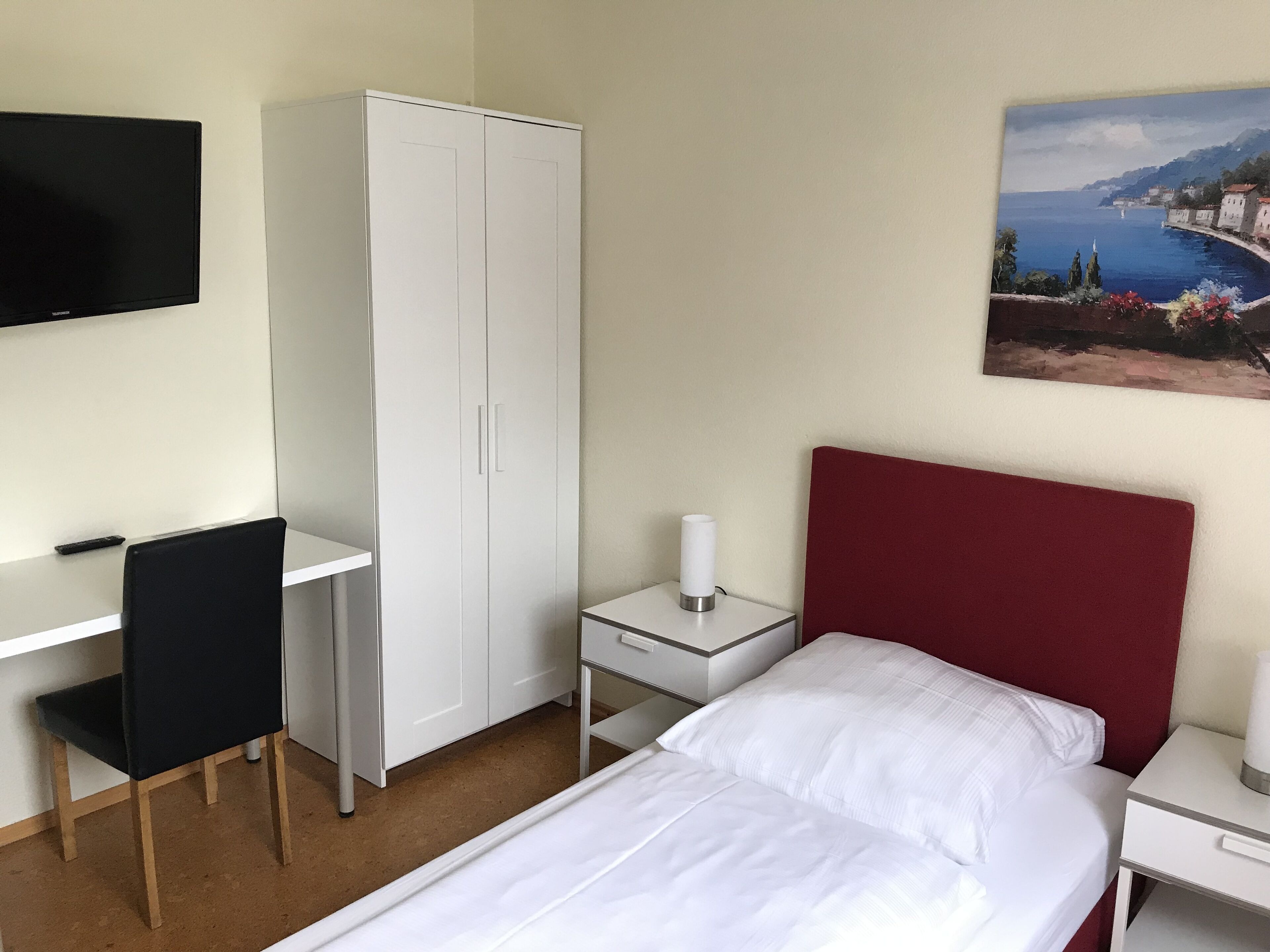 A hotel room with a bed, a desk, a wardrobe, a TV, and a painting of a coastal scene.