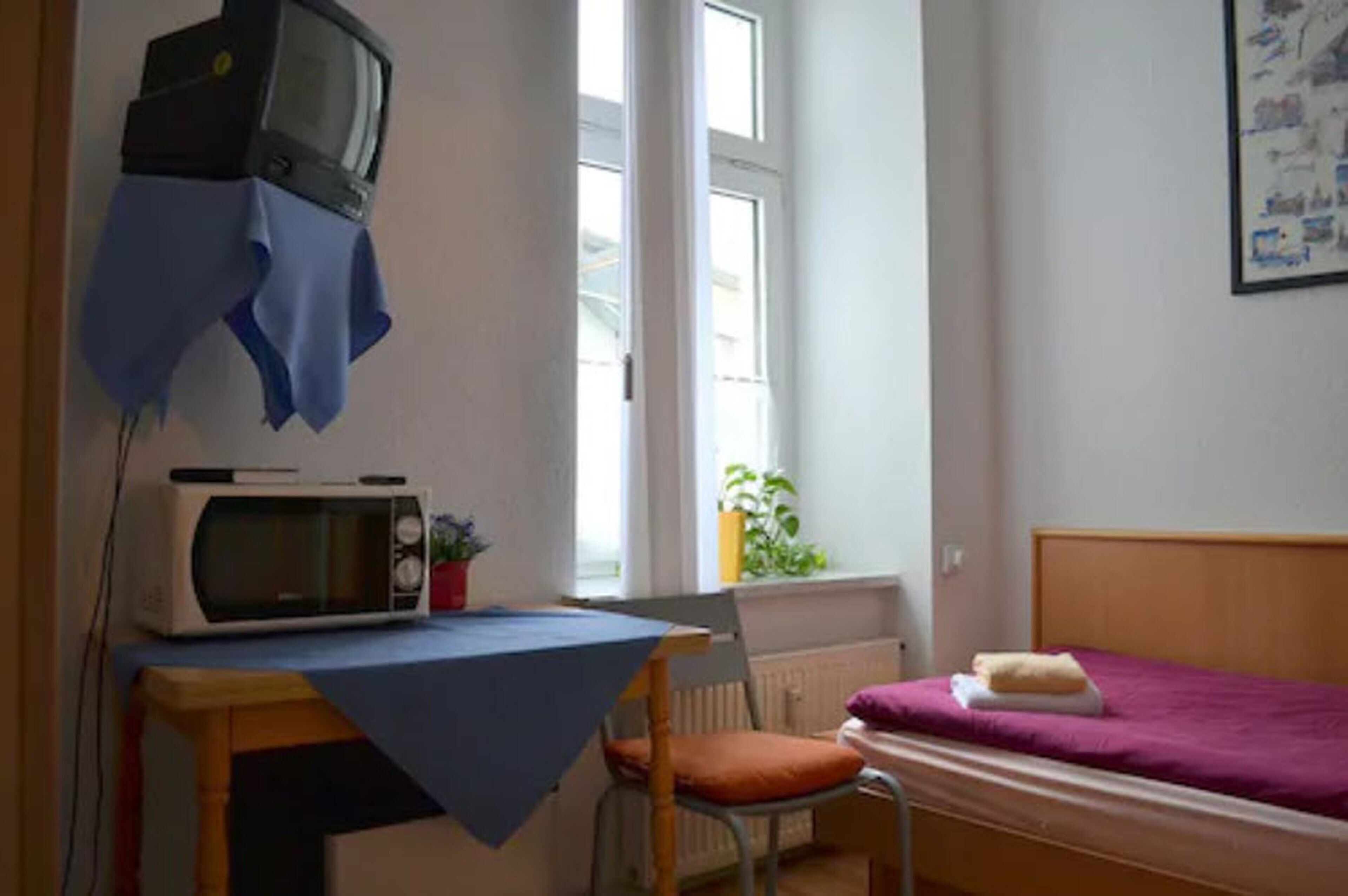 A small room with a bed, a table with a microwave, a chair, and a television on the wall.