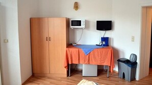 A small room with a wooden wardrobe, a table with an orange tablecloth, a microwave, a television, and a trash bin. - Pension Dessauer Hof (Halle (Saale))