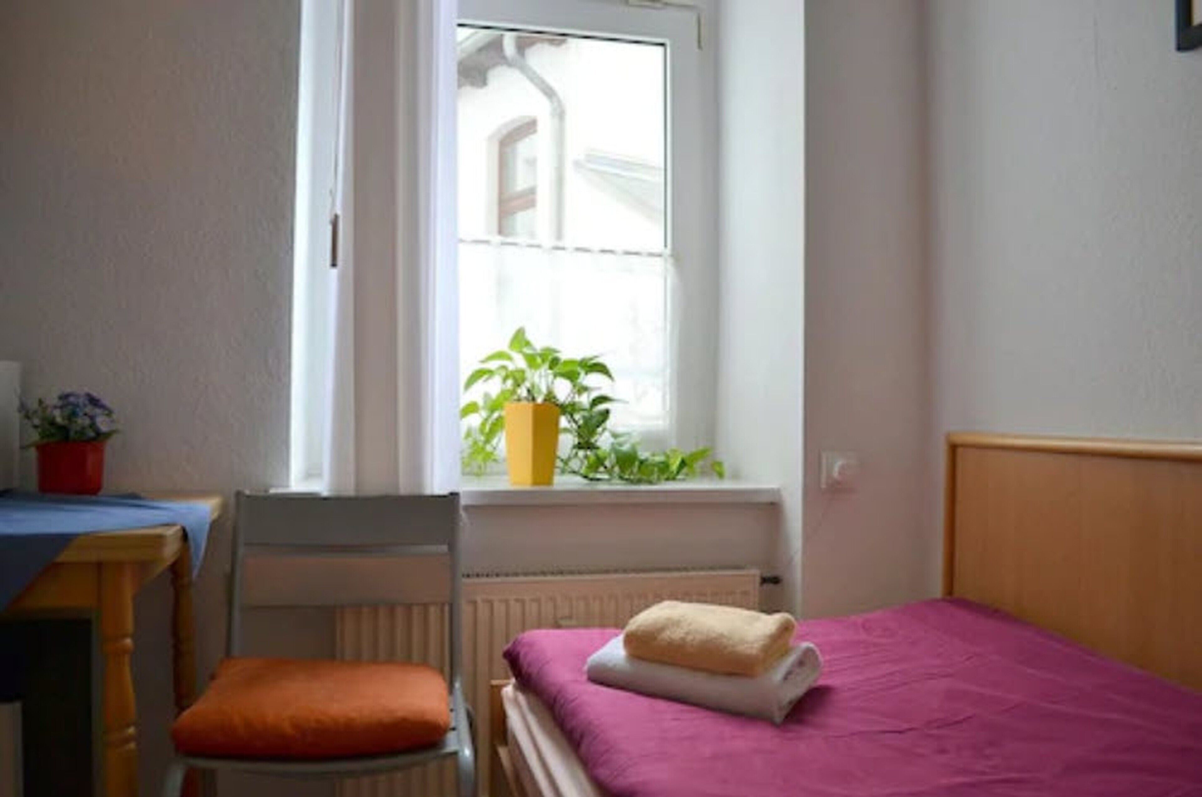 Una habitación pequeña y limpia con una cama individual, una silla, una mesa y ventana con vista a un edificio.