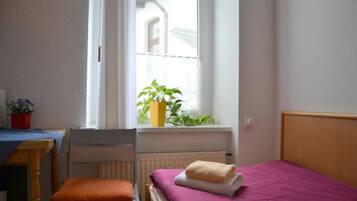 Una habitación pequeña y limpia con una cama individual, una silla, una mesa y ventana con vista a un edificio.