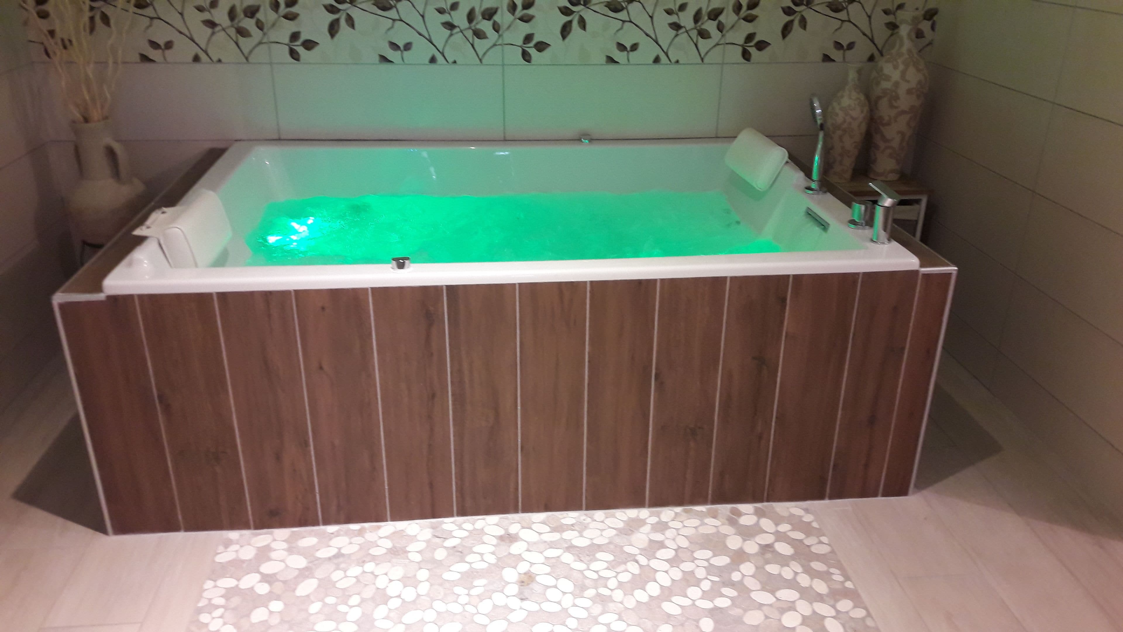 Una bañera de hidromasaje rectangular con revestimiento de madera y parte interior verde, ubicada en un baño con pared estampada y suelo de baldosas.