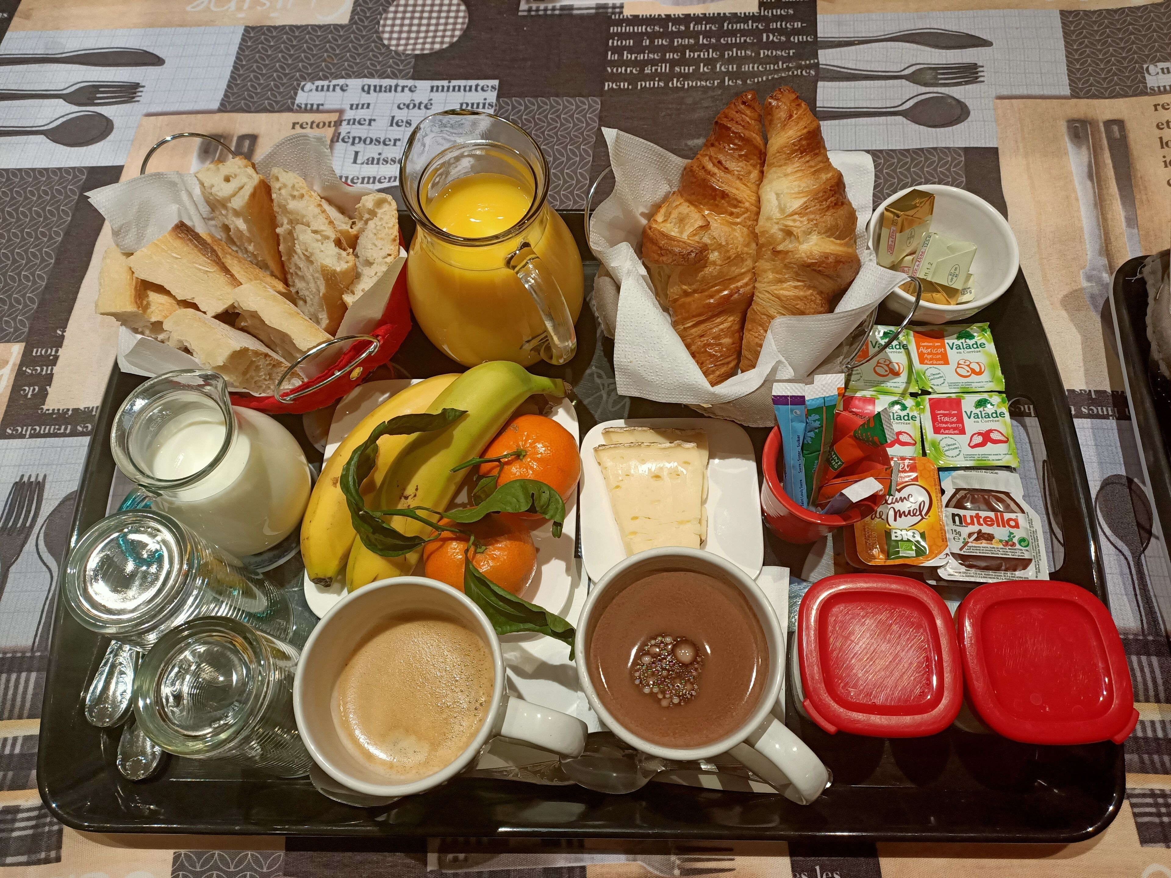En bakke med morgenmad, blandt andet croissanter, brød, bananer, appelsiner, ost, mælk, kaffe, Nutella og forskellige pålæg.