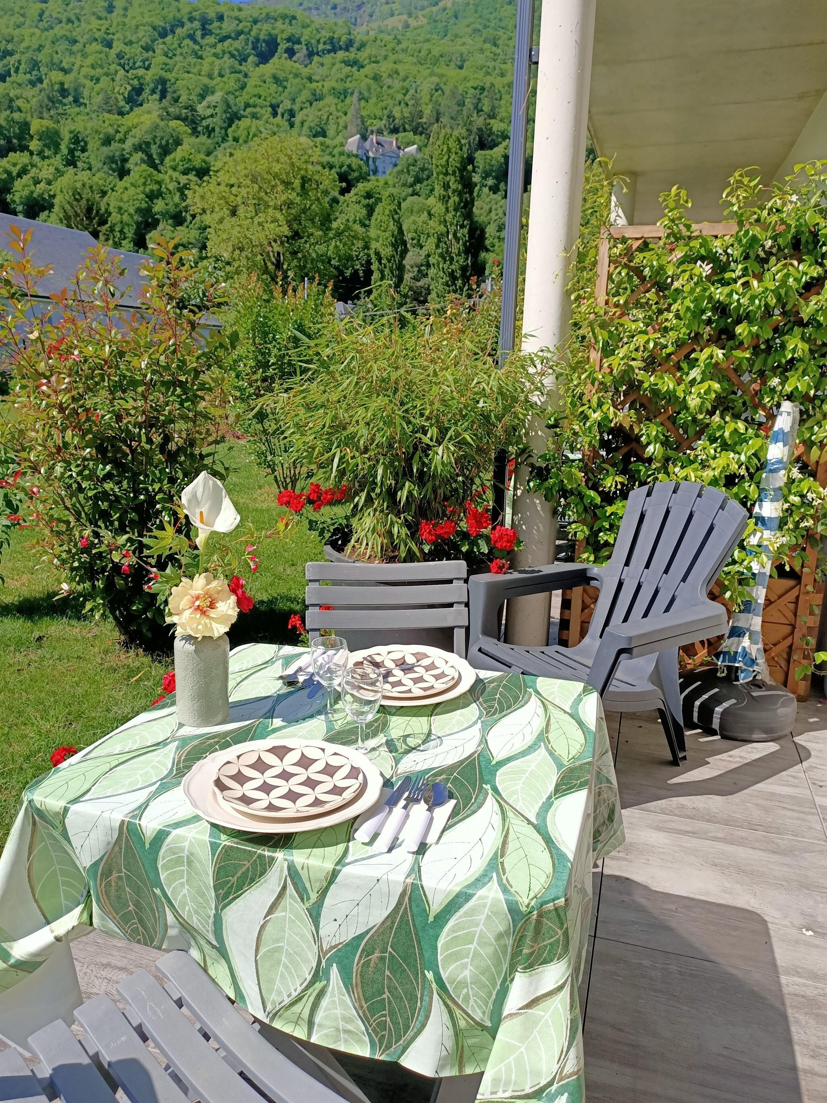 Ein Essbereich im Freien mit einem Tisch für zwei Personen, umgeben von Grünpflanzen und Blick auf ein bewaldetes Gebiet.