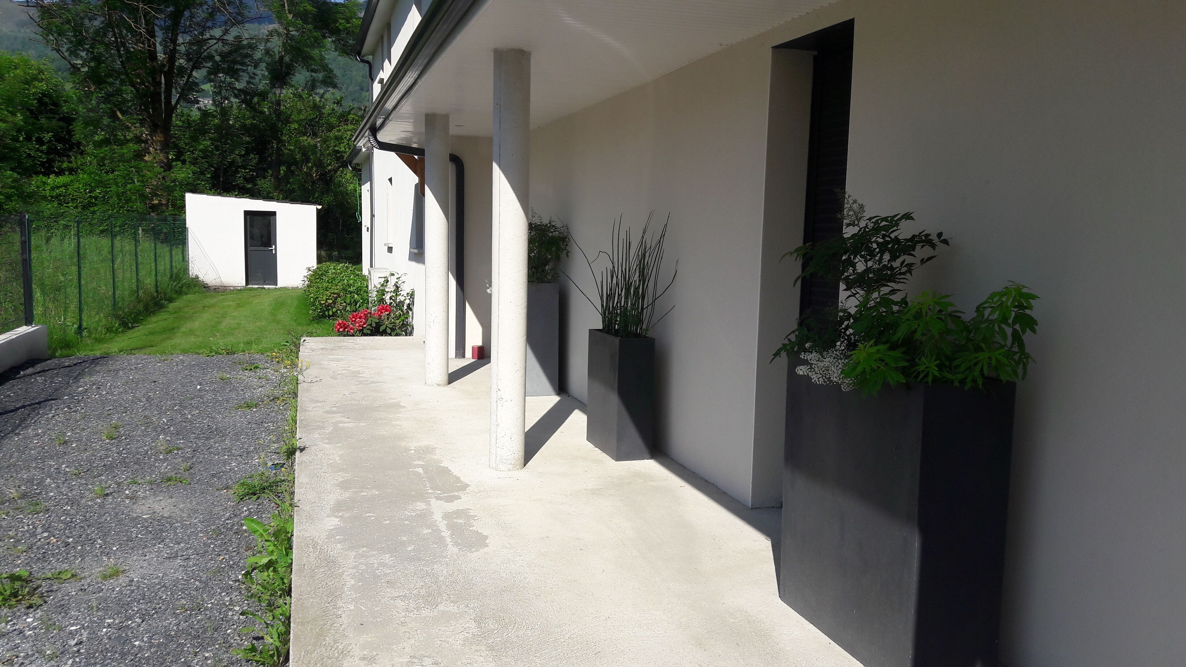 Un bâtiment moderne avec un chemin en béton menant à une entrée abritée. On peut apercevoir des plantes en pot et un petit espace aménagé en jardin.
