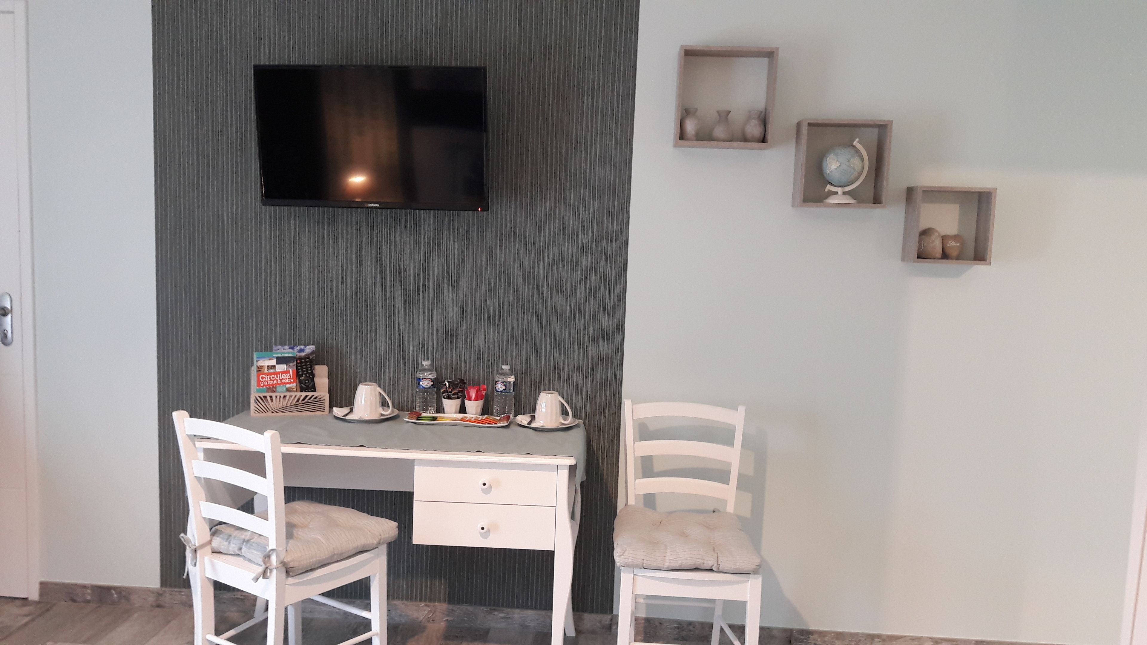 Una habitación con un televisor montado en una pared oscura, un escritorio con sillas y una mesita con una tetera, tazas y una botella de agua.