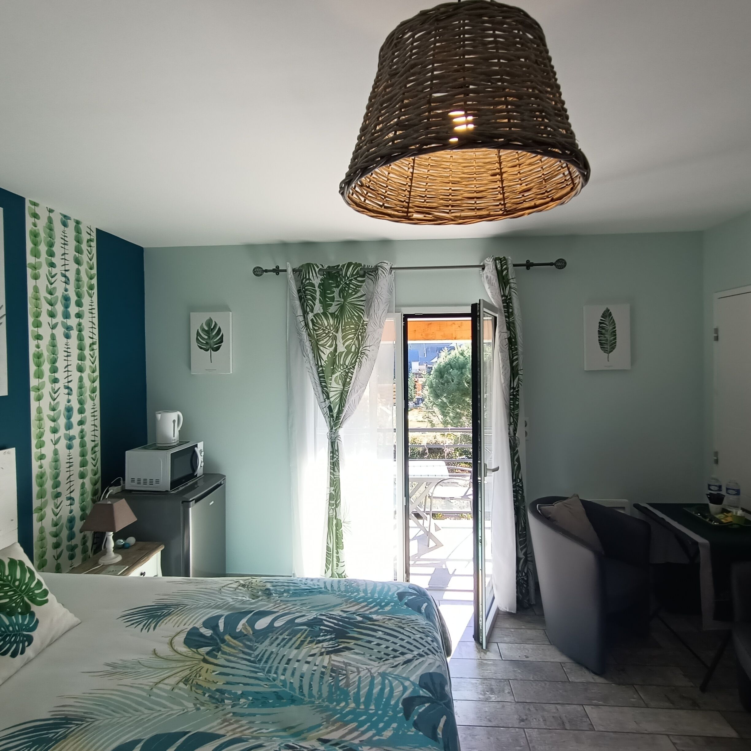 A bedroom with a bed, a desk, a chair, a hanging light fixture, and a view of the outside.