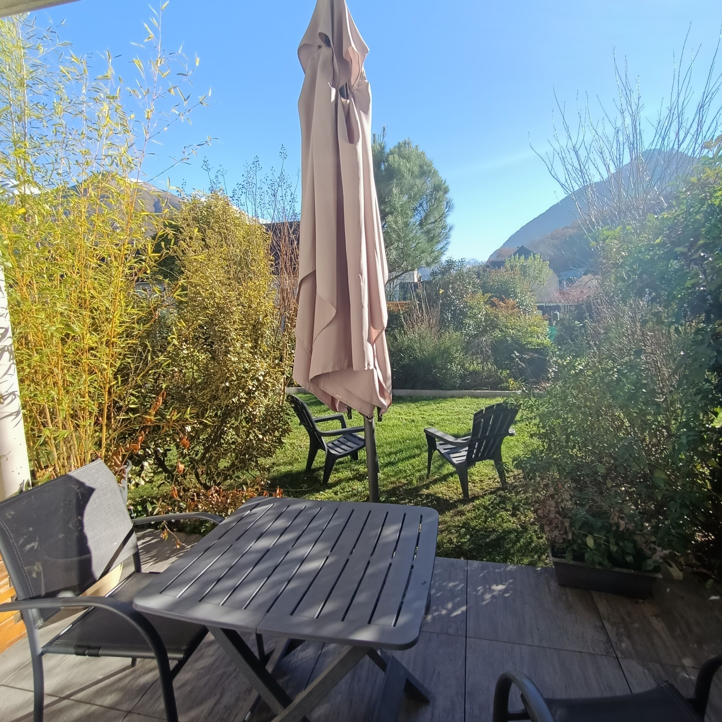 An outdoor patio with a table and chairs, surrounded by greenery and a clear sky.