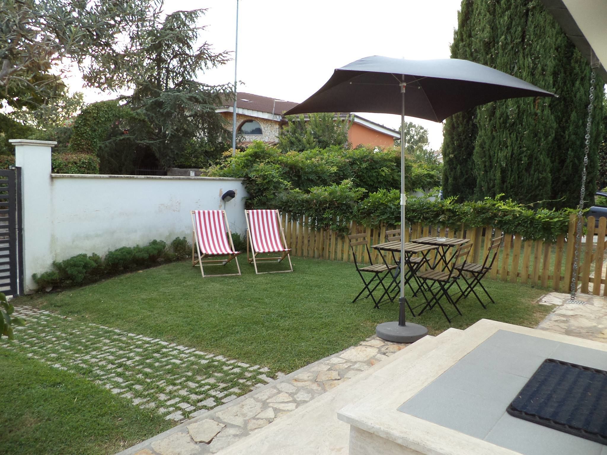 Un patio trasero con césped, dos sillones reclinables a rayas, una mesa con sillas y un paraguas grande.