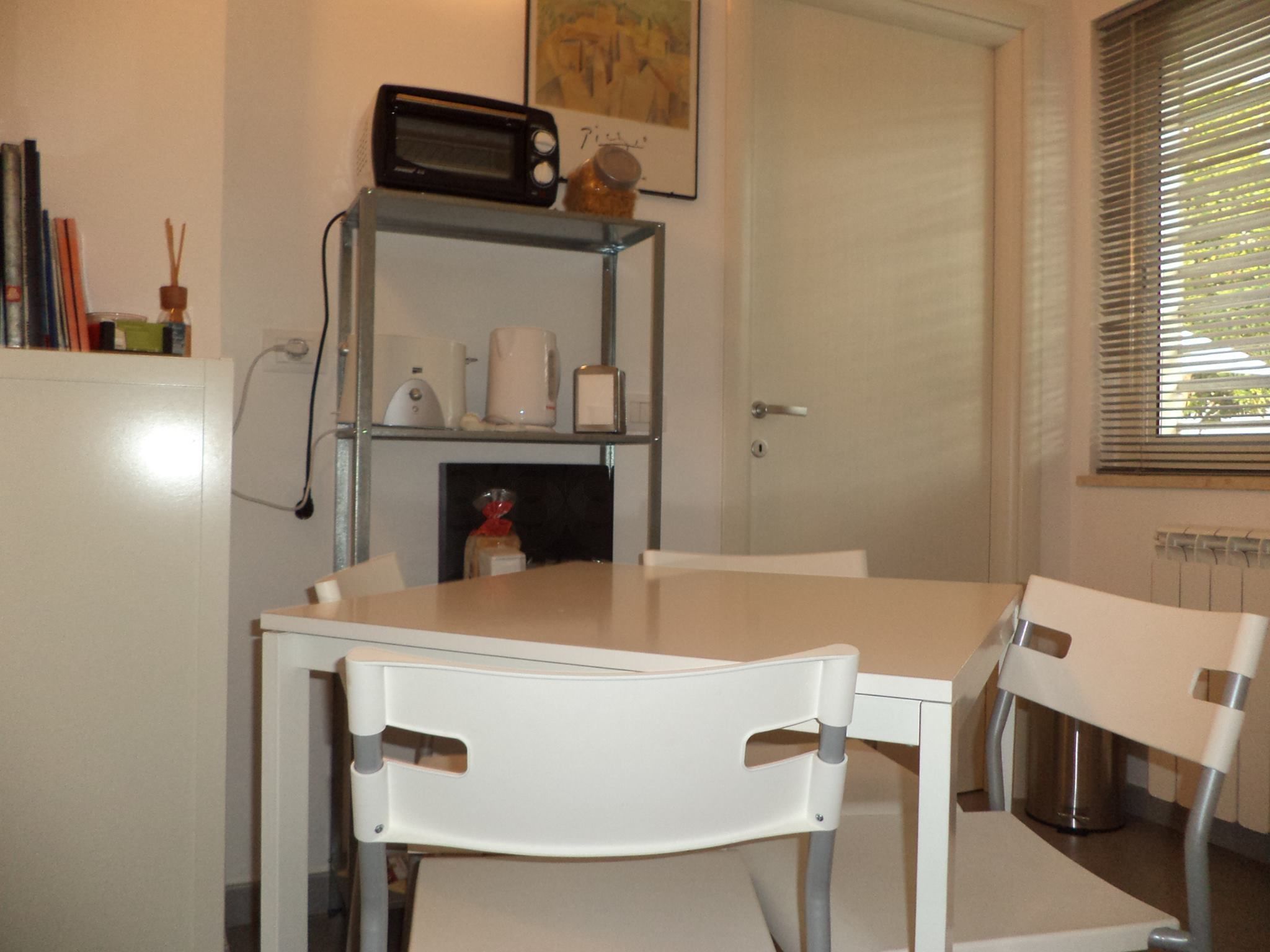 Una cocina pequeña con una mesa y sillas blancas, un tostador y una estantería con varios objetos.