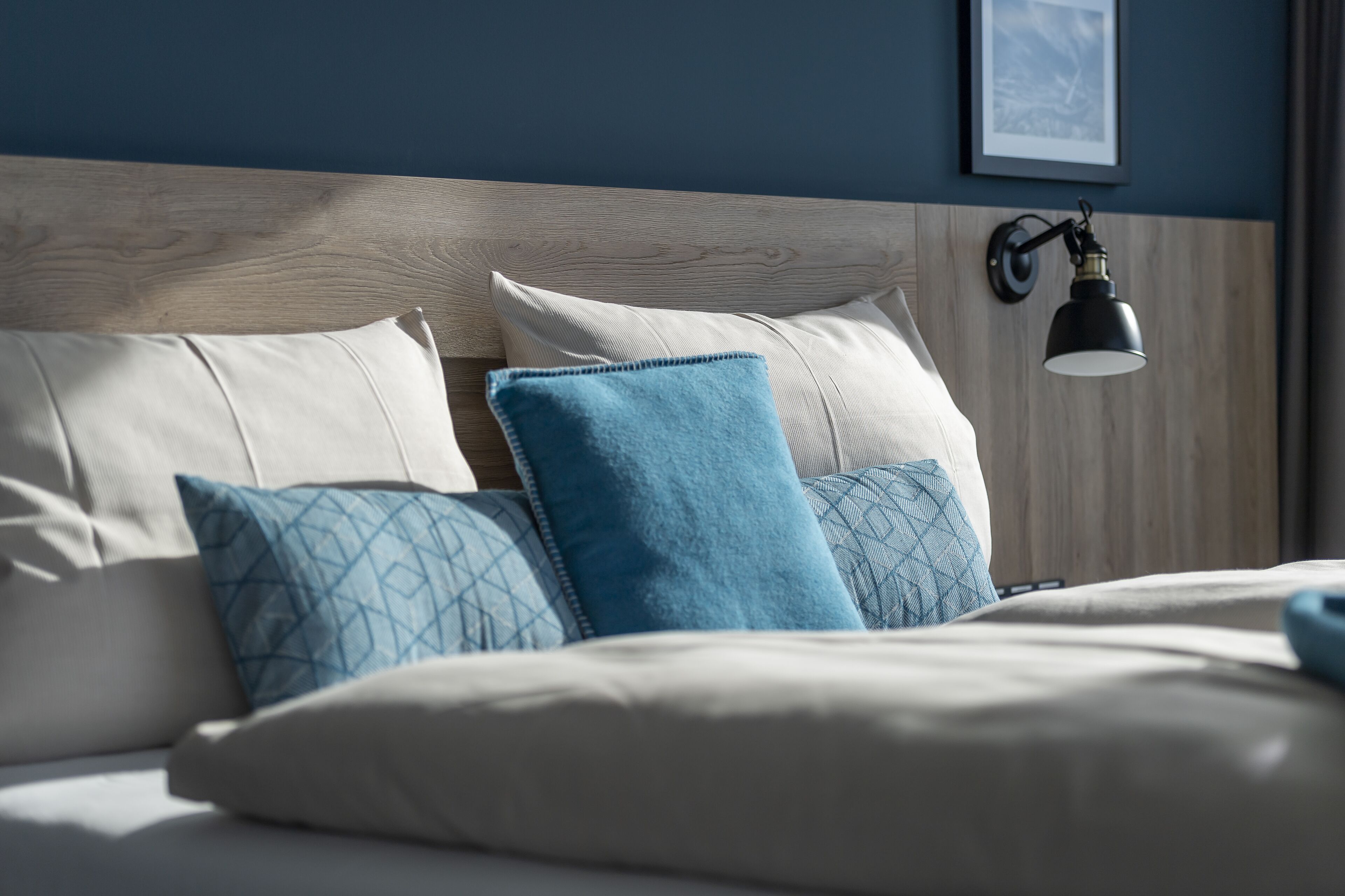A neatly made bed with a wooden headboard, multiple pillows, and a wall-mounted lamp.
