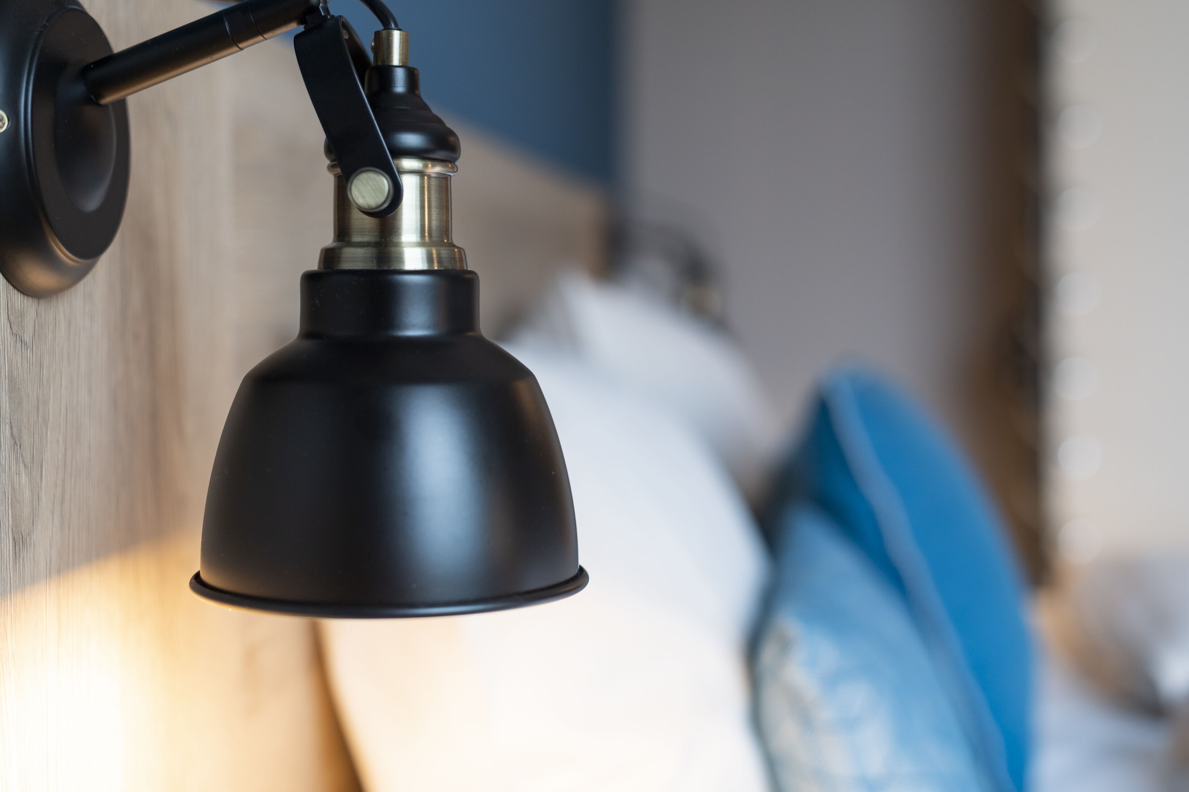 A wall-mounted lamp with a metallic base and a black shade, positioned above a bed with white and blue bedding.