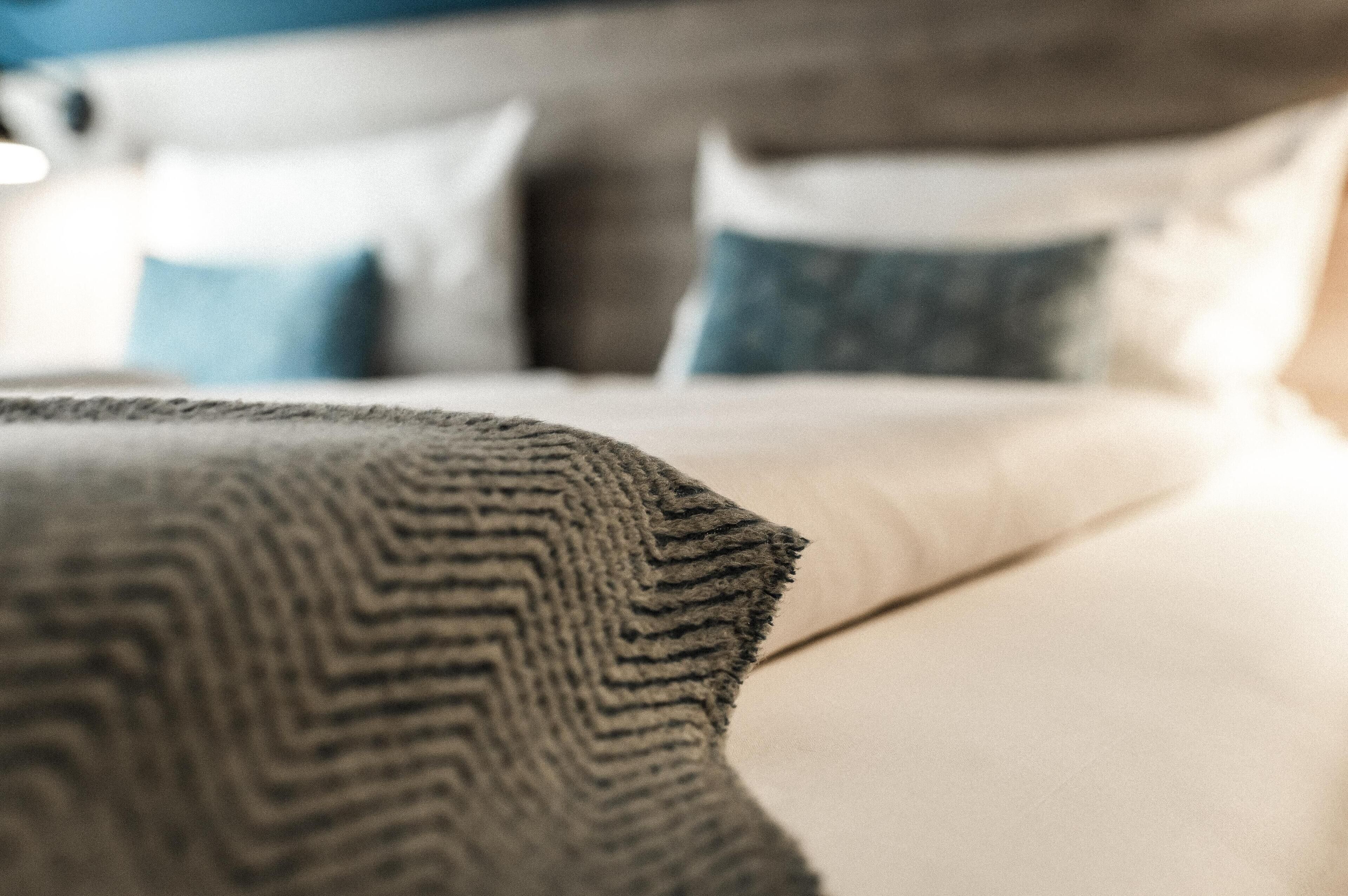 A close-up of a textured blanket on a bed with a neatly made bed and pillows in the background.