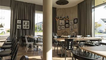 Un interior de restaurante con mesas y sillas de madera, cuadros en la pared y ventanas amplias con cortinas.