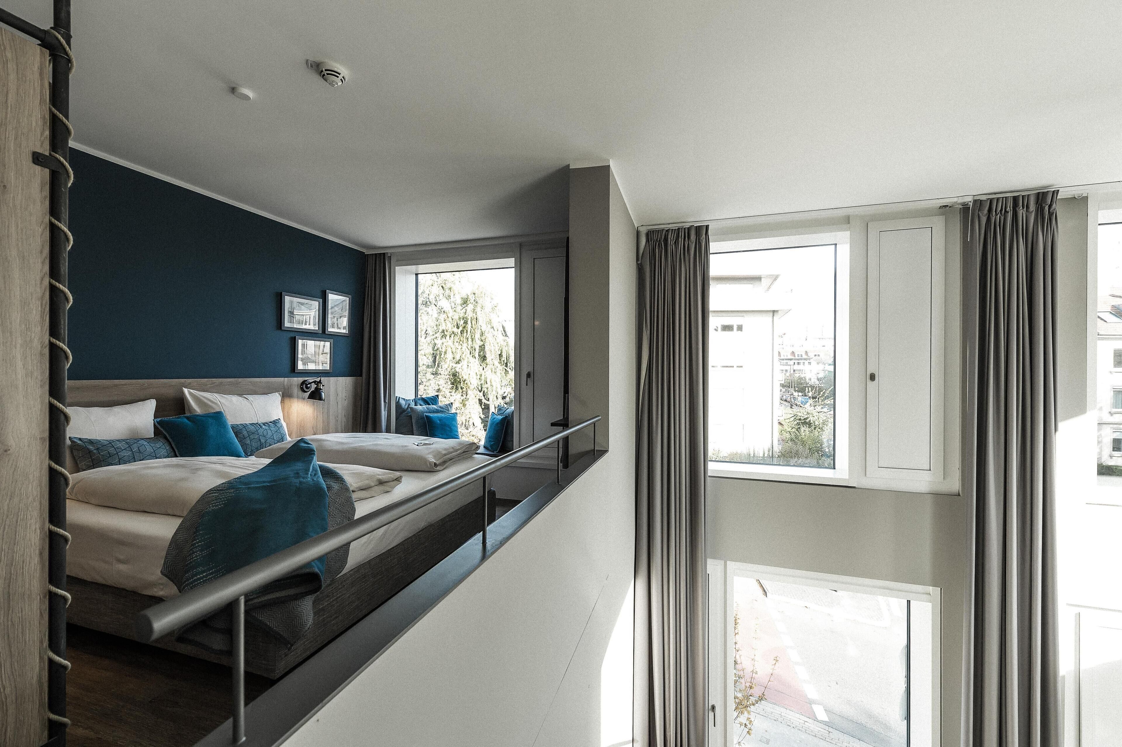 Una habitación de hotel moderna con balcón, cama con ropa de cama azul y blanca, y vista a la ciudad.