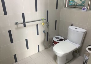 Un baño con inodoro blanco, ducha y secador de manos. Se ve una ventana con vista a un edificio y un estante pequeño con artículos de aseo.