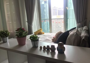 Un dormitorio con cama, almohadas, una mesita de noche con plantas y una ventana con cortinas.