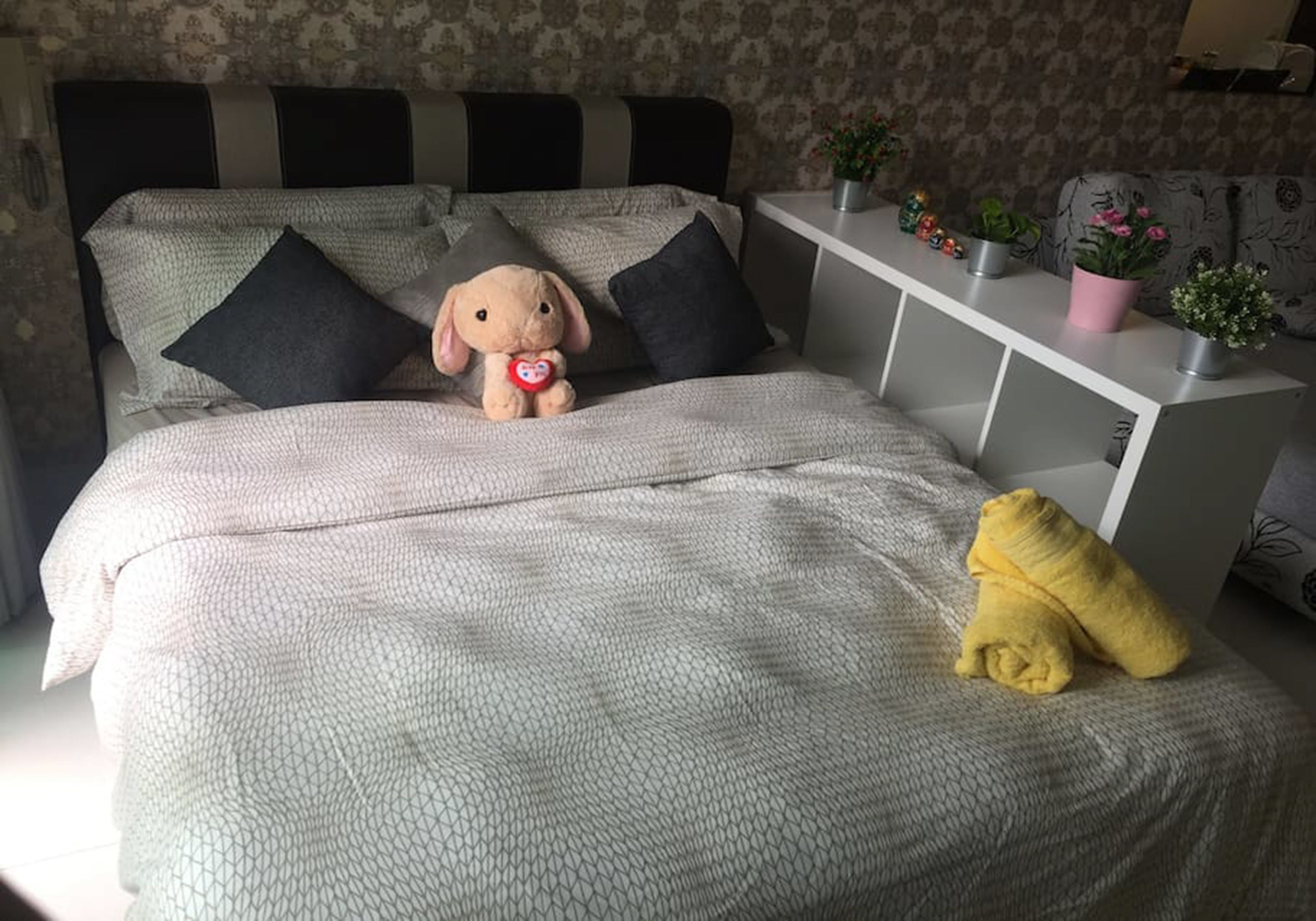 Un dormitorio con cama, almohadas, un peluche y una mesita de noche con plantas.