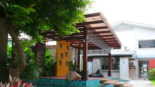 Uma área de estar ao ar livre coberta, com chão de azulejos, rodeada por vegetação e um edifício ao fundo.