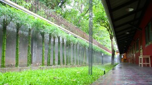 Uma galeria coberta com piso estampado, plantas de bambu no lado esquerdo e um edifício vermelho com janelas à direita.