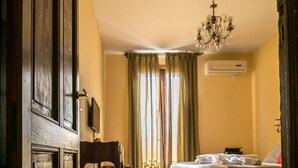 A hotel room with a wooden floor, a bed, a desk with a chair, a chandelier, and a window with curtains.