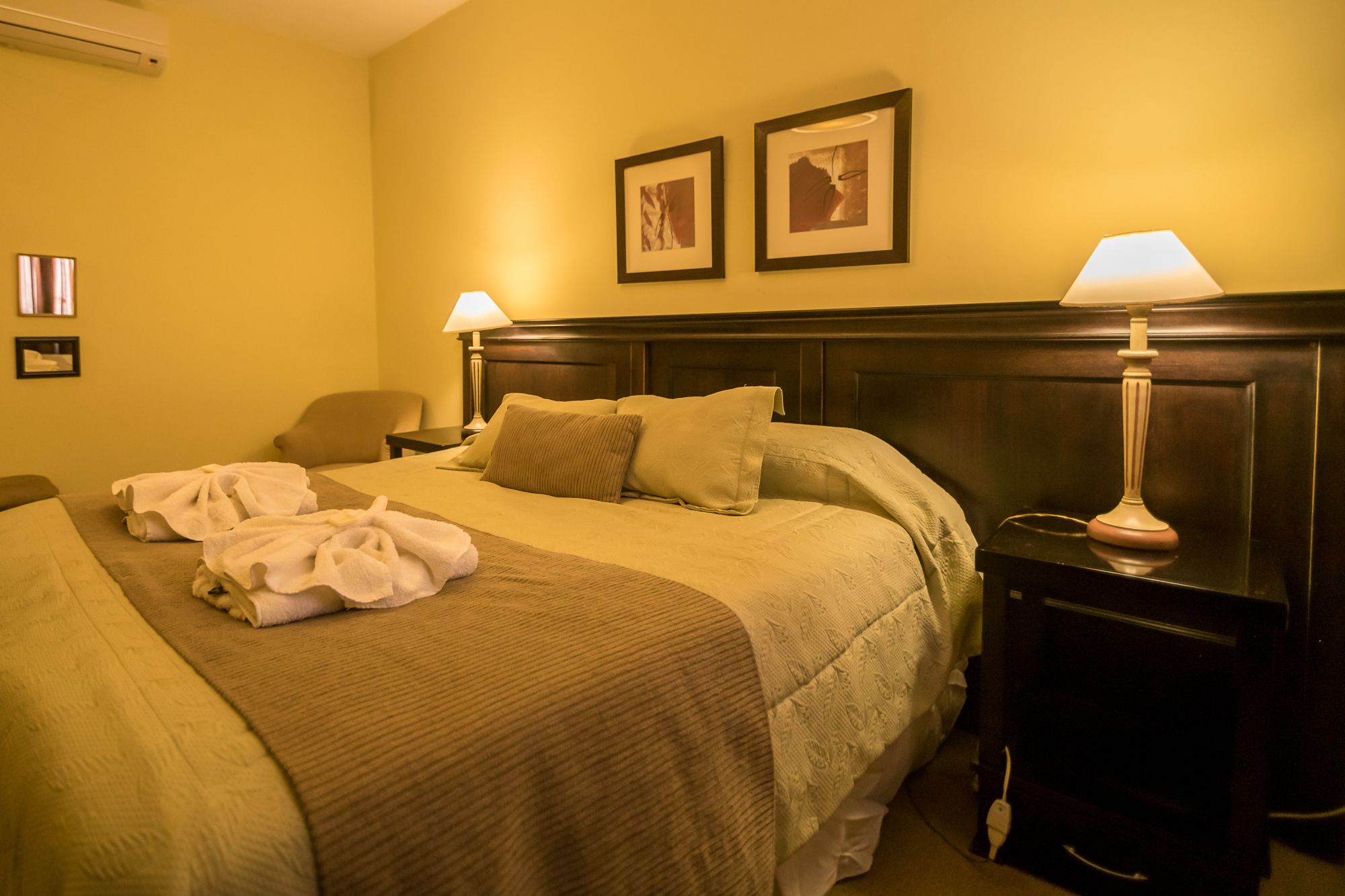 A hotel room with a large bed, two bedside lamps, a wooden headboard, and two framed pictures on the wall.