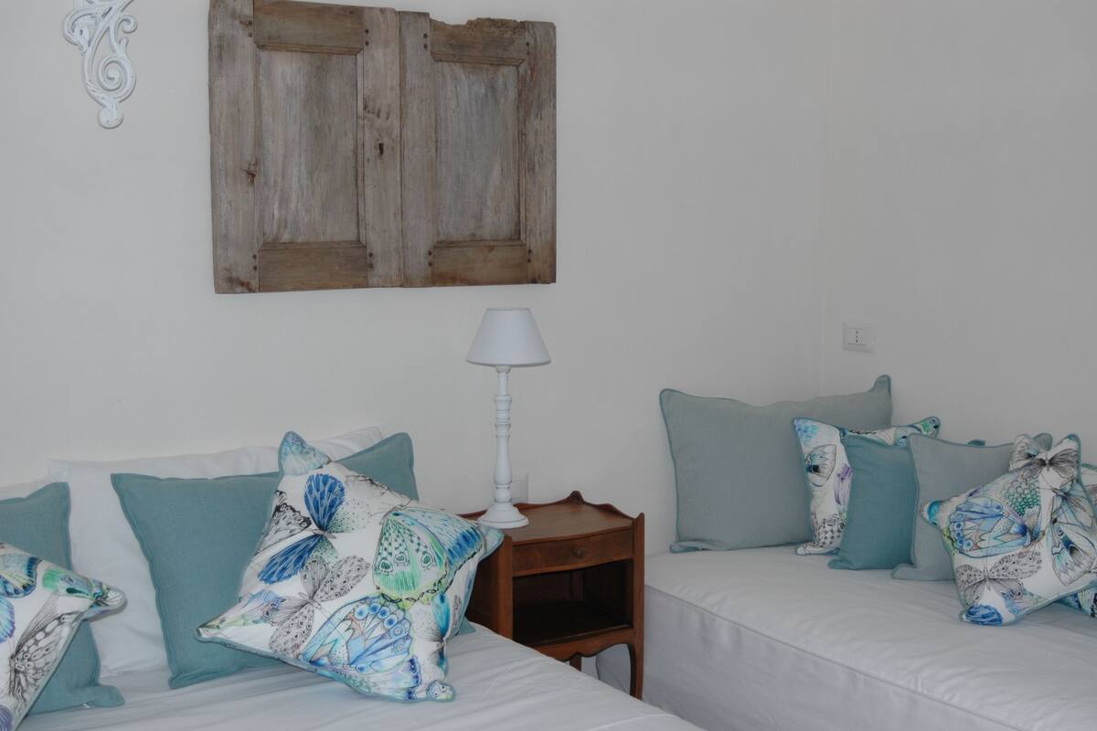A bedroom with two beds, each with teal and white bedding, a wooden headboard, a white wall, and a white lamp on a wooden nightstand.