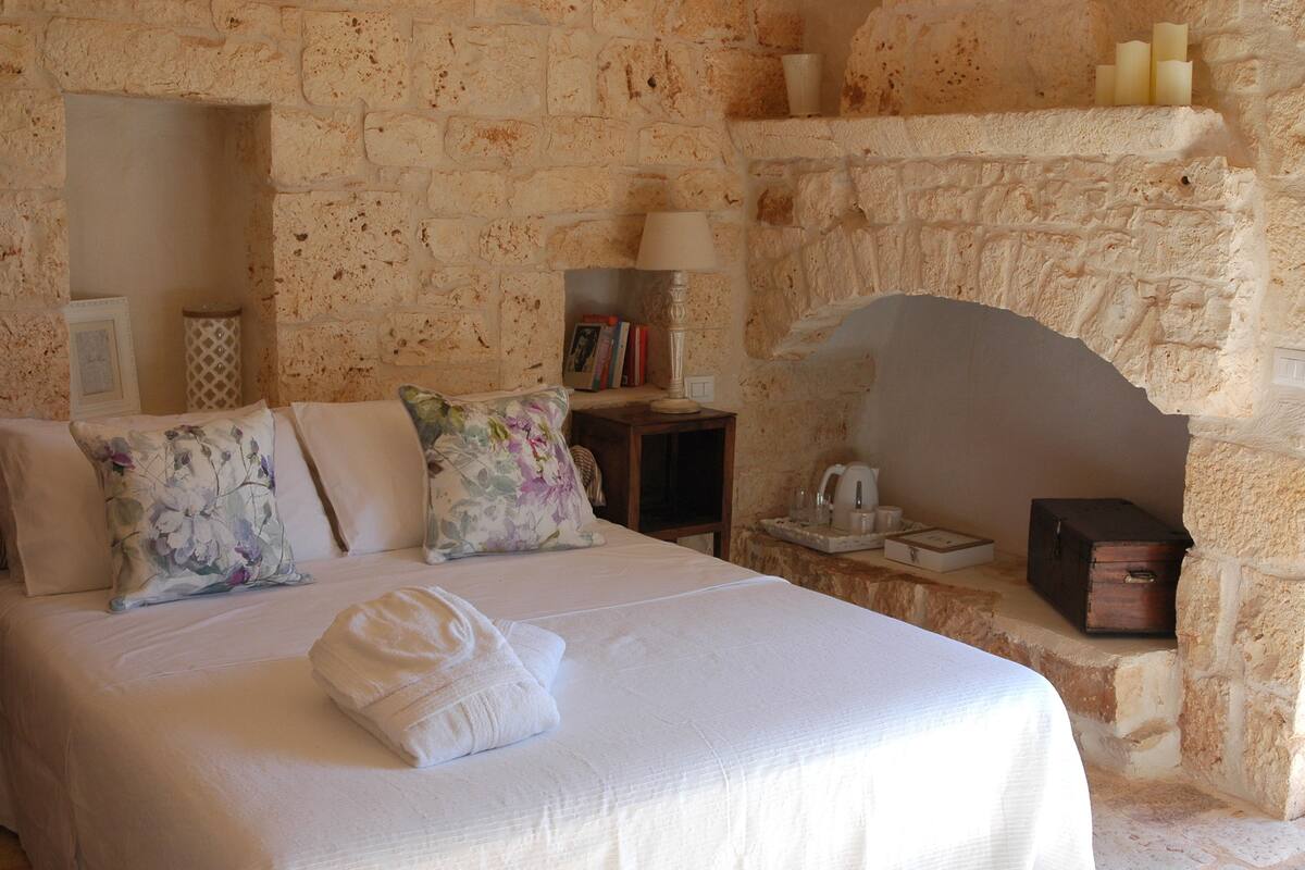 A bedroom with a stone wall, a bed with floral pillows, a bedside table with a lamp, and a small shelf with candles.