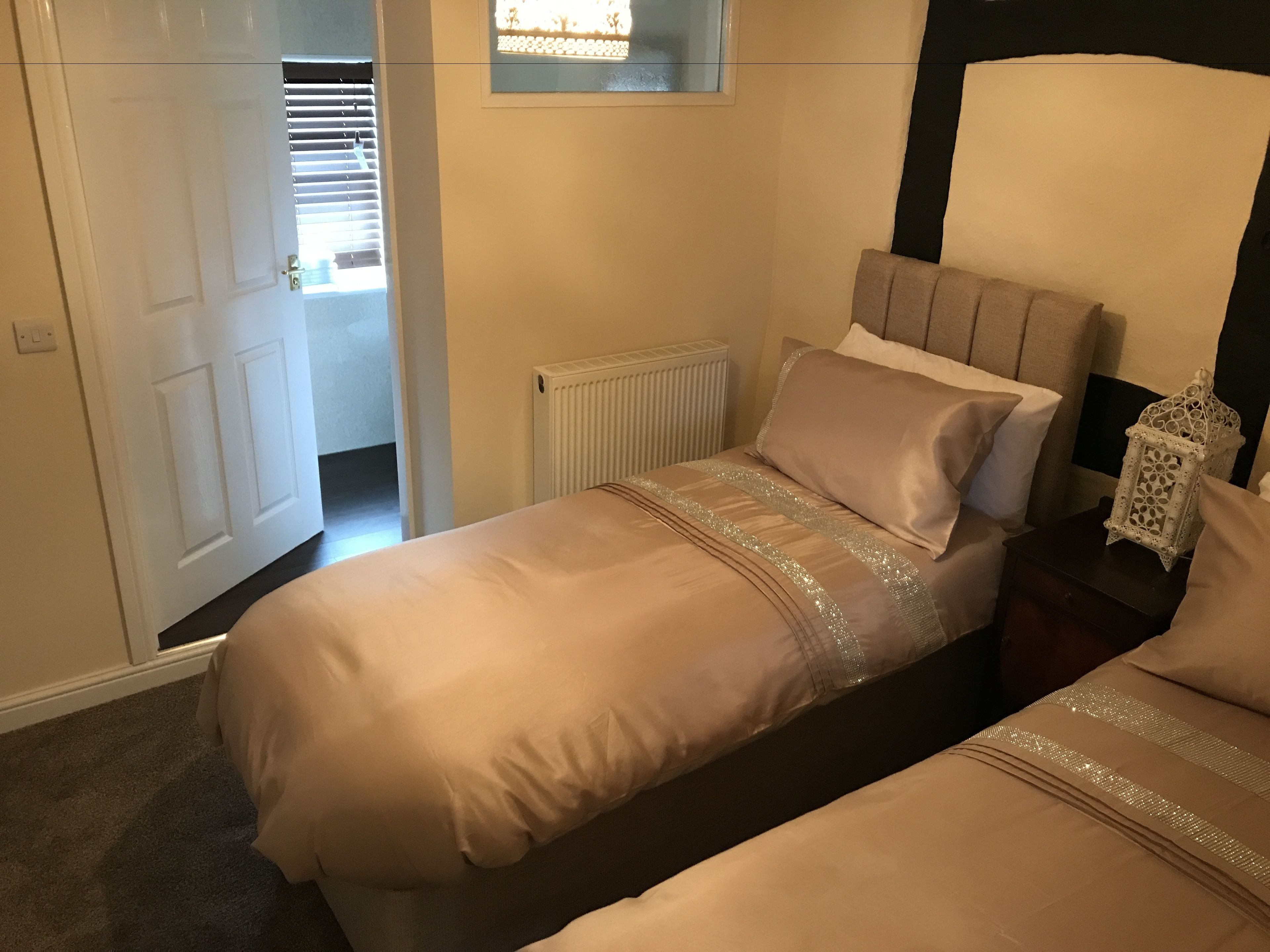 A bedroom with two beds, a radiator, a window with blinds, and a nightstand with a decorative lantern.