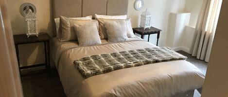 A neatly made bed with a patterned throw, a headboard, and multiple pillows. There are bedside tables with decorative items and wall-mounted mirrors.