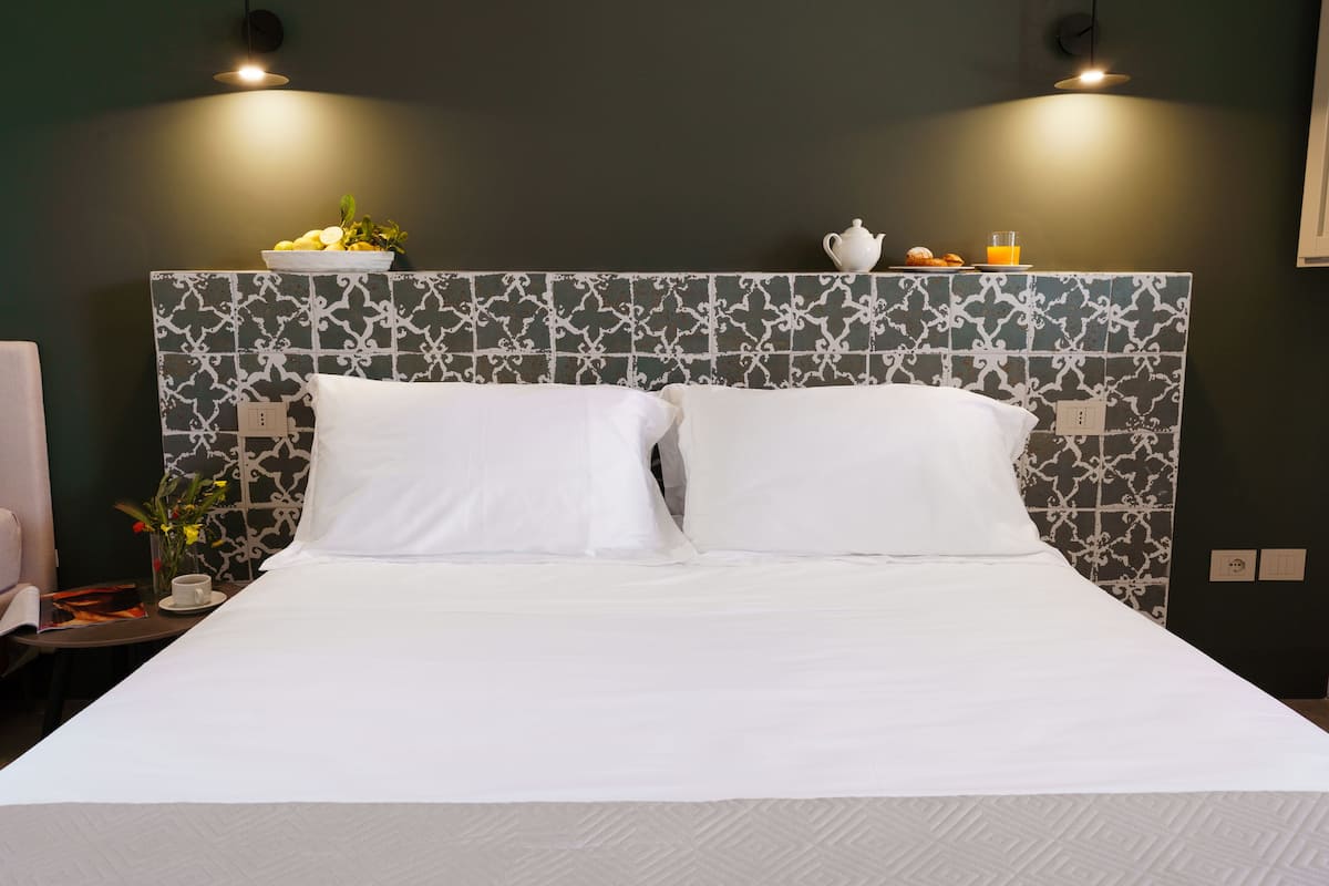 Un lit bien fait, avec du linge de lit blanc, un mur décoratif orné de motifs géométriques, et une petite table sur laquelle se trouvent une tasse et un vase de fleurs.