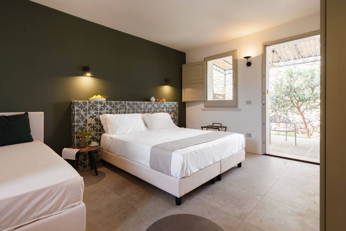 Une chambre moderne avec un grand lit, des tables de chevet, une petite table avec un vase, et une vue sur l’extérieur grâce à une porte vitrée.