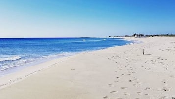 Une plage de sable avec des empreintes menant à l’eau, un ciel dégagé et un bâtiment visible au loin.