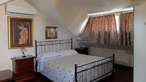 Un dormitorio con techo inclinado, cama con colcha estampada, cómoda de madera, mesita de noche, cuadro enmarco y ventana con cortinas florales.