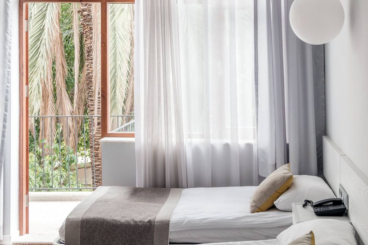 Ein modernes Hotelzimmer mit zwei Betten, einem großen Fenster mit durchsichtigen Vorhängen und Blick auf Palmen.
