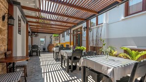 Een terras buiten met tafels en stoelen, houten balken erboven en potplanten.