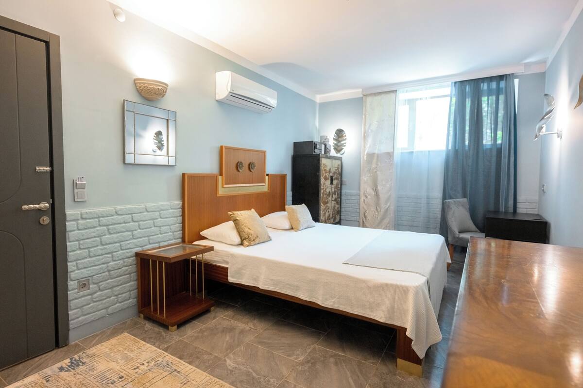 Ein Hotelzimmer mit einem Bett, einem Nachttisch aus Holz, einem kleinen Tisch, einem Sessel und einem Fenster mit Vorhängen.