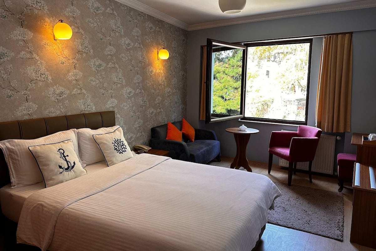 A hotel room with a large bed, two wall-mounted lamps, a window with a view of greenery, a small round table, and a red chair.