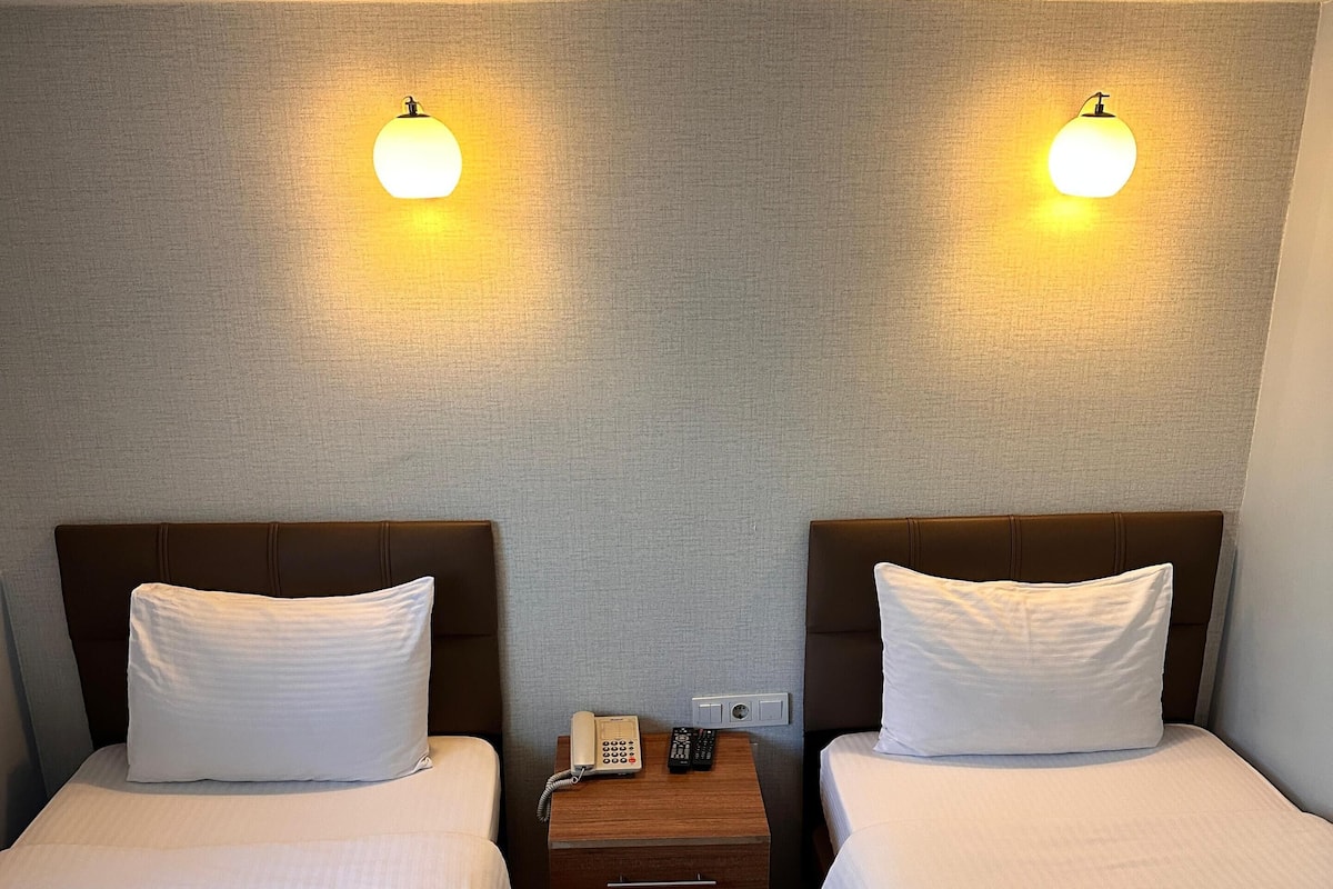 A hotel room with two single beds, a wooden nightstand with a phone and remote, and wall-mounted lights.