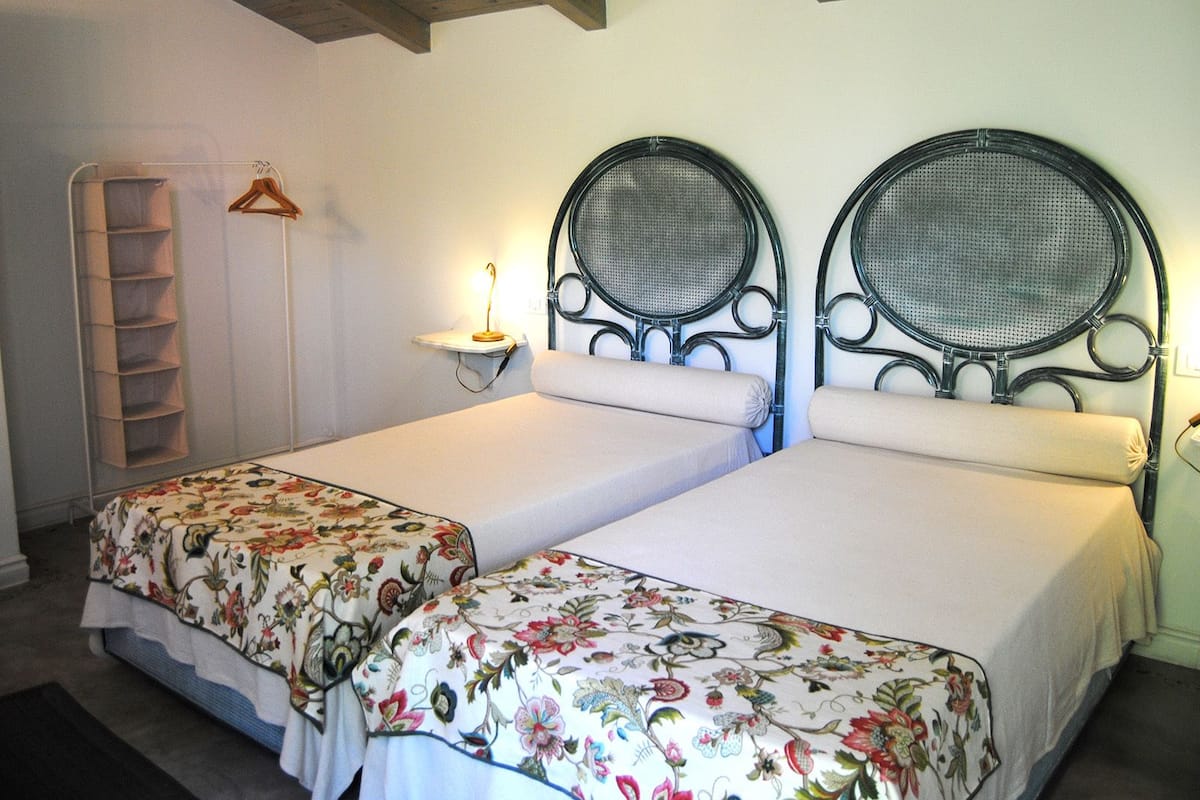 A room with two single beds, a bedside table with a lamp, a wooden shelf unit, and a wall-mounted shelf.