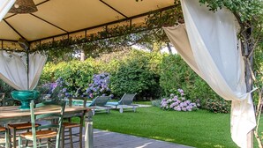 An outdoor patio with a table and chairs, surrounded by lush greenery and flowering plants. - Villa Hermes (San Ferdinando)