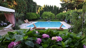 A swimming pool with two lounge chairs and a covered area, surrounded by lush greenery and flowering plants. - Villa Hermes (San Ferdinando)