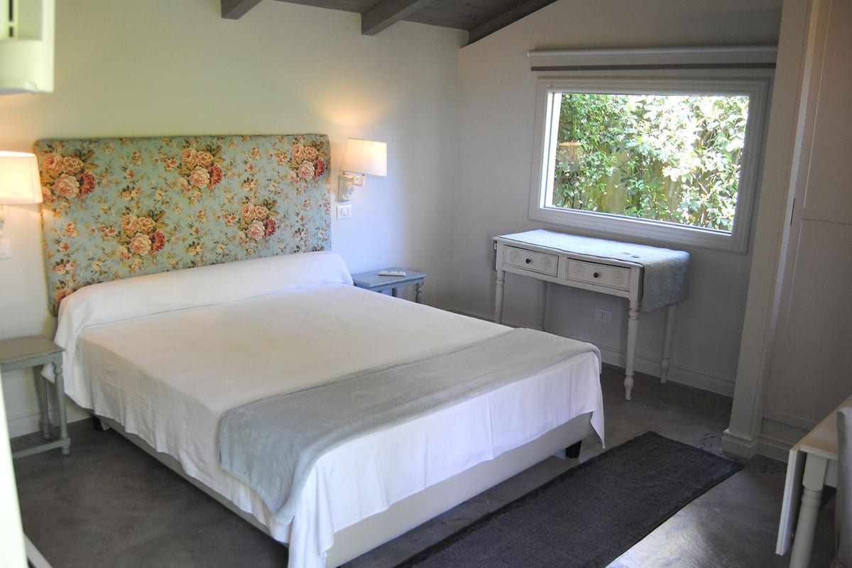 A bedroom with a bed, a desk, a lamp, and a window with a view of greenery.
